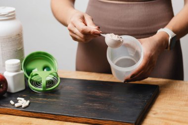 Spor giysilerinde elinde kaşık olan atletik bir kadın, beyaz amino asit kapsülleri, muz ve elmalı, protein içeceği yapan beyaz protein tozunun bir kısmını ahşap masaya koyar..