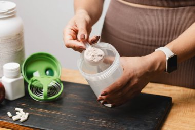 Spor giysilerinde elinde kaşık olan atletik bir kadın, beyaz amino asit kapsülleriyle ahşap masadaki süt tozu tozunun bir kısmına protein içeceği koyuyor..