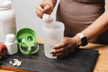 Spor giysilerinde elinde kaşık olan atletik bir kadın, beyaz amino asit kapsülleriyle ahşap masadaki süt tozu tozunun bir kısmına protein içeceği koyuyor..