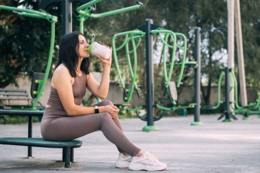 Spor kıyafetleri içinde sportif güzel bir kadın. Spor sahasında spor yaptıktan sonra, bir içecekten protein içeceği ve spor beslenmesi içiyor..
