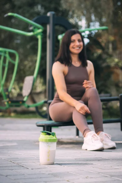 Shaker with sport nutrition protein drink on a sports ground and athletic pretty woman in sportswear after outdoors workout on a background.