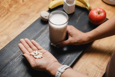 Spor kıyafetleri içindeki atletik kadın elinde bir bardak protein içeceği kokteyli ve masada beyaz amino asit kapsülleri ile protein tozu ve meyve ölçme kaşığı tutuyor..