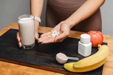 Spor kıyafetleri içindeki atletik kadın elinde bir bardak protein içeceği kokteyli ve masada beyaz amino asit kapsülleri ile protein tozu ve meyve ölçme kaşığı tutuyor..