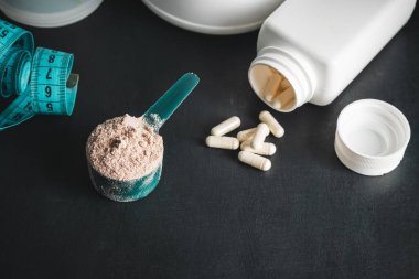 Ölçme kaşığında çikolatalı süt tozu, beyaz amino asit kapsülleri, vitaminler ve kreatin ölçüm bandı, koyu arkaplanda plastik karıştırıcı. Vücut geliştirme gıda takviyeleri.