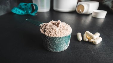 Ölçme kaşığında çikolatalı süt tozu, beyaz amino asit kapsülleri, vitaminler ve kreatin ölçüm bandı, koyu arkaplanda plastik karıştırıcı. Vücut geliştirme gıda takviyeleri.