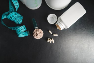 Ölçme kaşığında çikolatalı süt tozu, beyaz amino asit kapsülleri, vitaminler ve kreatin, mezura, koyu arkaplanda titretici. Vücut geliştirme gıda malzemeleri, üst görünüm..