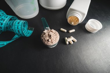 Ölçme kaşığında çikolatalı süt tozu, beyaz amino asit kapsülleri, vitaminler ve kreatin ölçüm bandı, koyu arkaplanda plastik karıştırıcı. Vücut geliştirme gıda takviyeleri.