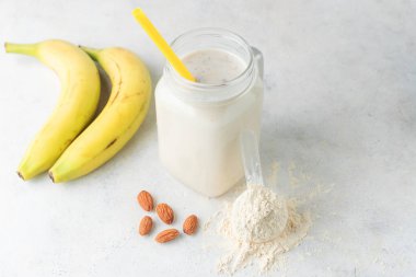 Cam kavanoz protein milkshake ya da smoothie ve beyaz arka planda kaşık, muz ve badem fıstığı içinde beyaz protein tozu. Spor beslenmesi, vücut geliştirme gıda takviyeleri..