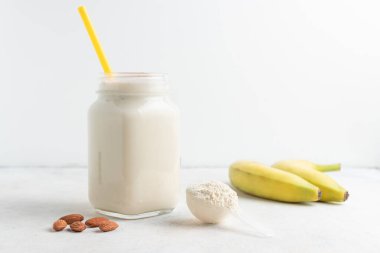 Cam kavanoz protein milkshake ya da smoothie ve beyaz arka planda kaşık, muz ve badem fıstığı içinde beyaz protein tozu. Spor beslenmesi, vücut geliştirme gıda takviyeleri..