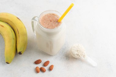 Cam kavanoz protein milkshake ya da smoothie ve beyaz arka planda kaşık, muz ve badem fıstığı içinde beyaz protein tozu. Spor beslenmesi, vücut geliştirme gıda takviyeleri..