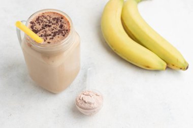 Cam kavanoz çikolatalı milkshake ya da smoothie ve beyaz arka planda muzlar ölçme kaşığında beyaz protein tozu. Spor beslenmesi, vücut geliştirme gıda takviyeleri..