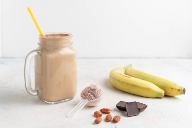 Cam kavanoz protein milkshake ya da smoothie ve beyaz zemin üzerinde kaşık, muz, çikolata küpleri ve badem fıstığı içeren beyaz protein tozu. Spor beslenmesi, vücut geliştirme gıda takviyeleri..