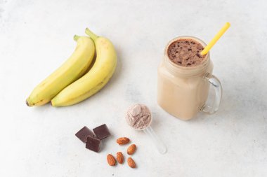 Cam kavanoz protein milkshake ya da smoothie ve beyaz zemin üzerinde kaşık, muz, çikolata küpleri ve badem fıstığı içeren beyaz protein tozu. Spor beslenmesi, vücut geliştirme gıda takviyeleri..