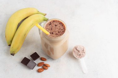 Cam kavanoz protein milkshake ya da smoothie ve beyaz zemin üzerinde kaşık, muz, çikolata küpleri ve badem fıstığı içeren beyaz protein tozu. Spor beslenmesi, vücut geliştirme gıda takviyeleri..