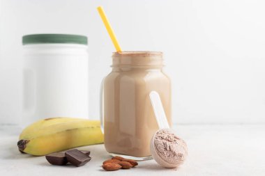 Ölçme kaşığında çikolatalı süt tozu, beyaz zemin üzerinde cam kavanoz protein milkshake içeceği veya smoothie, muz, çikolata küpleri ve badem. Spor besinleri, gıda takviyeleri.