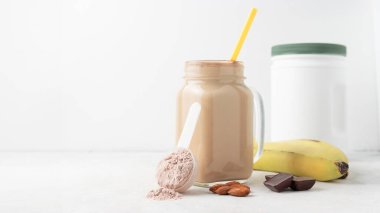 Ölçme kaşığında çikolatalı süt tozu, beyaz zemin üzerinde cam kavanoz protein milkshake içeceği veya smoothie, muz, çikolata küpleri ve badem. Spor besinleri, gıda takviyeleri.