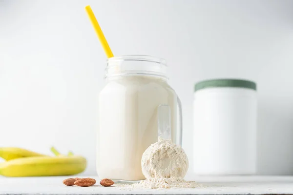 Whey protein powder in measuring spoon, glass jar of protein milkshake drink or smoothie, bananas and almond nuts on a white background. sport nutrition, bodybuilding food supplements.