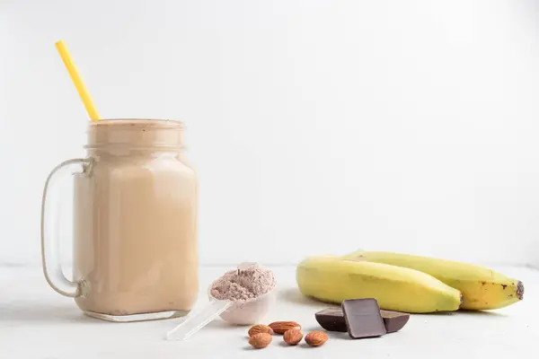 Glass jar of protein milkshake drink or smoothie and whey protein powder in measuring spoon, bananas, chocolate cubes, almond nuts on white background. sport nutrition, bodybuilding food supplements.
