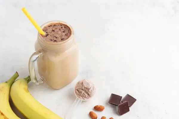 Glass jar of protein milkshake drink or smoothie and whey protein powder in measuring spoon, bananas, chocolate cubes, almond nuts on white background. sport nutrition, bodybuilding food supplements.
