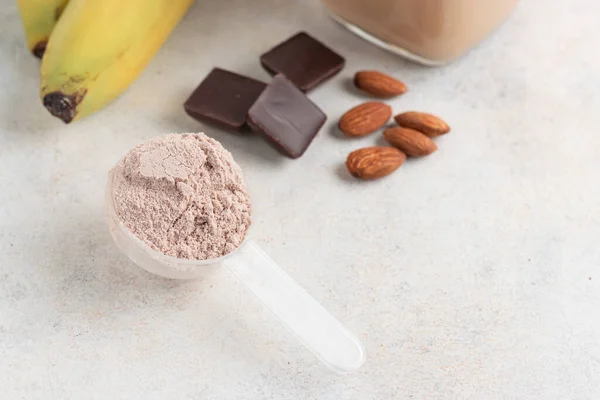 Chocolate whey protein powder in measuring spoon, glass jar of protein milkshake drink or smoothie, bananas, chocolate cubes and almonds on white background. sport nutrition, food supplements.