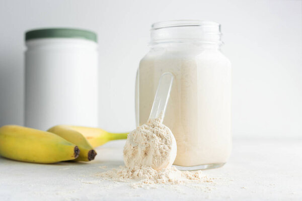 Glass jar of protein milkshake drink or smoothie and whey protein powder in measuring spoon, bananas on a white background. sport nutrition, bodybuilding food supplements.