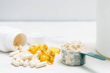 Beyaz kaşıkta beyaz protein tozu, beyaz amino asit kapsülleri, vitaminler ve sarı kapsüller beyaz arka planda Omega 3, sağlıklı beslenme ve vücut geliştirme besinleri..
