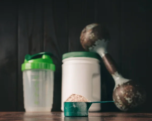 Chocolate whey protein powder in measuring spoon, old rusty dumbbell, shaker, plastic bottle on dark background. healthy eating, bodybuilding food supplements.