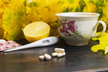 Kapsüller, haplar, su toplayan ilaçlar, termometre, ahşap bir masada limonlu çay fincanı ve sarı yapraklar. Sonbahar nezlesi, grip ve koronavirüs tedavisi kavramı