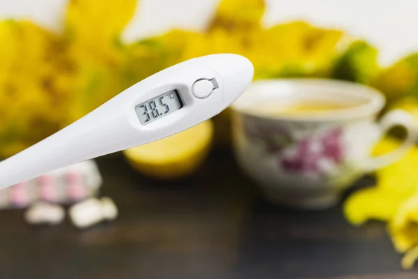 Digital thermometer indicates high temperature 38.5, above table with tea cup, lemon, capsules, pills and yellow leaves. Concept of treatment fever, autumn cold, flu and coronavirus.