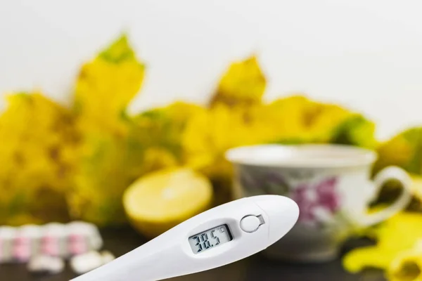 Digital thermometer indicates high temperature 38.5, above table with tea cup, lemon, capsules, pills and yellow leaves. Concept of treatment fever, autumn cold, flu and coronavirus.