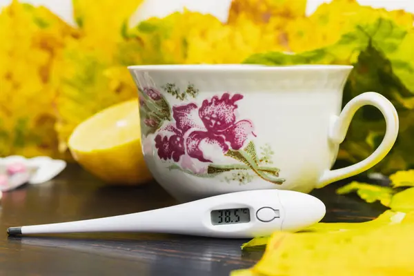 Digital thermometer indicates high temperature 38.5 on a table with tea cup, lemon, capsules, pills and yellow leaves. Concept of treatment fever, autumn cold, flu and coronavirus.