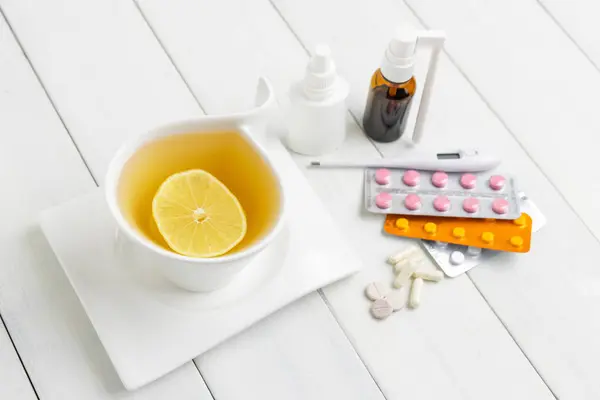 Tea cup with lemon, cold remedies, medicine, thermometer, capsules and pills in a blister pack on wooden table. Concept of home treatment flu and coronavirus.