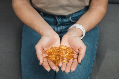 Genç kadın elinde omega-3 balık yağı, ellerinde vitamin sarısı kapsüller, evdeki kanepede oturuyor, yakın çekim manzarası.