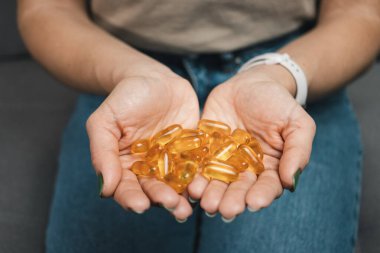 Genç kadın elinde omega-3 balık yağı, ellerinde vitamin sarısı kapsüller, evdeki kanepede oturuyor, yakın çekim manzarası.