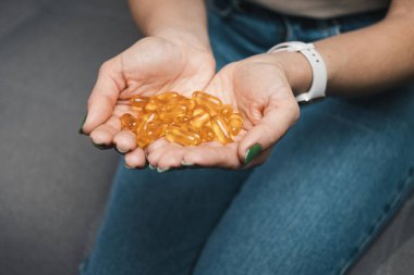Genç kadın elinde omega-3 balık yağı, ellerinde vitamin sarısı kapsüller, evdeki kanepede oturuyor, yakın çekim manzarası.