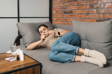 Ateşli üzgün kadın evdeki kanepede uzanıyor ve sıcaklığını dijital termometreyle kontrol ediyor. Soğuk algınlığı ilaçları, ilaçlar ve haplar masada. Mevsimsel hastalıklar, koronavirüs ve grip kavramı.