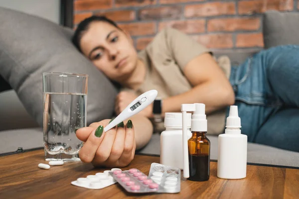 Sick woman with fever lying on sofa at home, holding digital thermometer showing high temperature of 38.1. cold remedies, medicine and pills on table. Concept of seasonal diseases, coronavirus and flu.