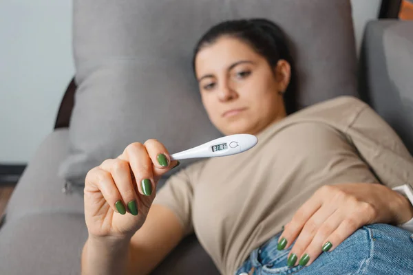 Woman with fever and headache is lying on the sofa at home and holding a digital thermometer showing a high temperature of 38.1. Concept of seasonal diseases such as coronavirus, cold and flu.