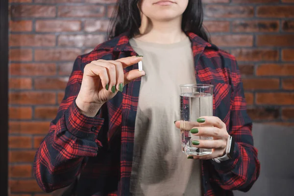 Young woman takes a medicine, white pill and holds in a hand glass of water, close-up view.