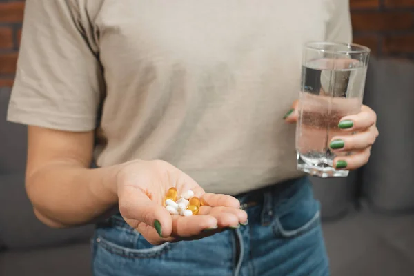 stock image Young woman holds various medical pills and capsules in a hand and glass of water, close-up view.