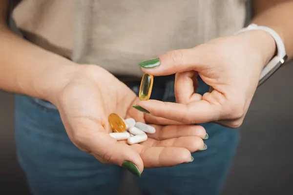 Young woman takes one capsule of omega 3 supplement and holds pile of various medical pills and capsules in a hand, close-up view.