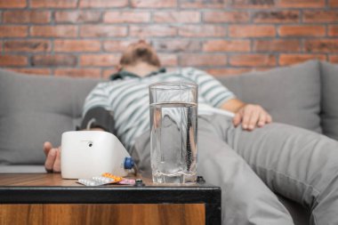 Hipertansiyonu ve baş ağrısı olan, evdeki koltukta oturan, elinde modern dijital tonometreyle kan basıncını ölçen, ön planda haplarla dolu su toplamaya odaklanan bir adam..