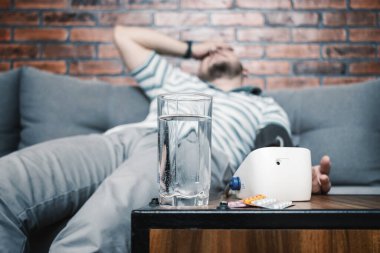 Hipertansiyonu ve baş ağrısı olan, evdeki koltukta oturan, elinde modern dijital tonometreyle kan basıncını ölçen, ön planda haplarla dolu su toplamaya odaklanan bir adam..