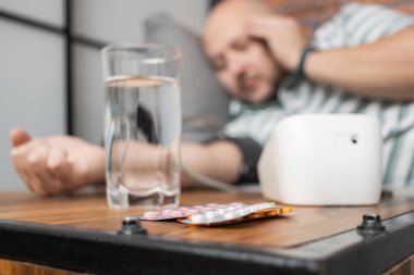 Hipertansiyonu ve baş ağrısı olan, evdeki koltukta uzanan, elinde modern dijital tonometreyle kan basıncını ölçen, ön planda haplarla dolu su toplamaya odaklanan bir adam..