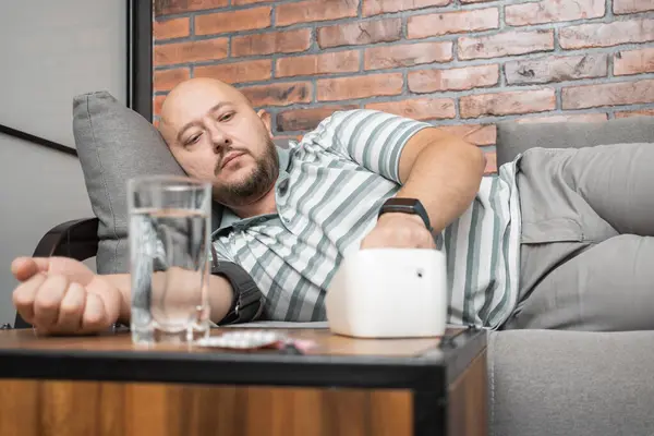 Young adult man lying on a sofa at home measures blood pressure on a hand with modern digital tonometer.