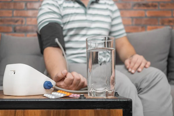 Defocused background with man sitting on sofa at home and measures blood pressure on hand with modern digital tonometer, focus on blister pack with pills on foreground.