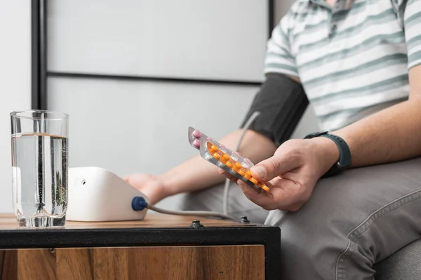 Unrecognizable man sitting on a sofa at home and measures blood pressure on a hand with modern digital tonometer and holding blister packs with pills, medicines .