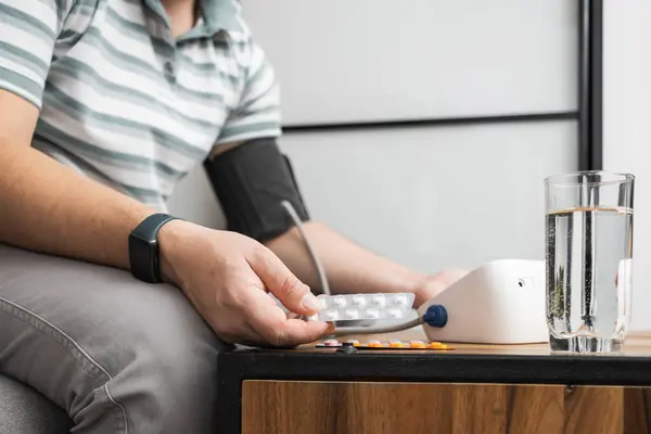 Unrecognizable man sitting on a sofa at home and measures blood pressure on a hand with modern digital tonometer and holding blister packs with pills, medicines .