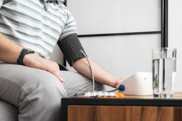 Unrecognizable man sitting on a sofa at home and measures blood pressure on a hand with modern digital tonometer.