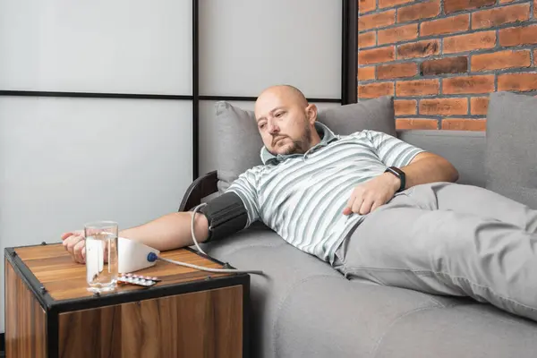 Young adult man lying on a sofa at home measures blood pressure on a hand with modern digital tonometer.